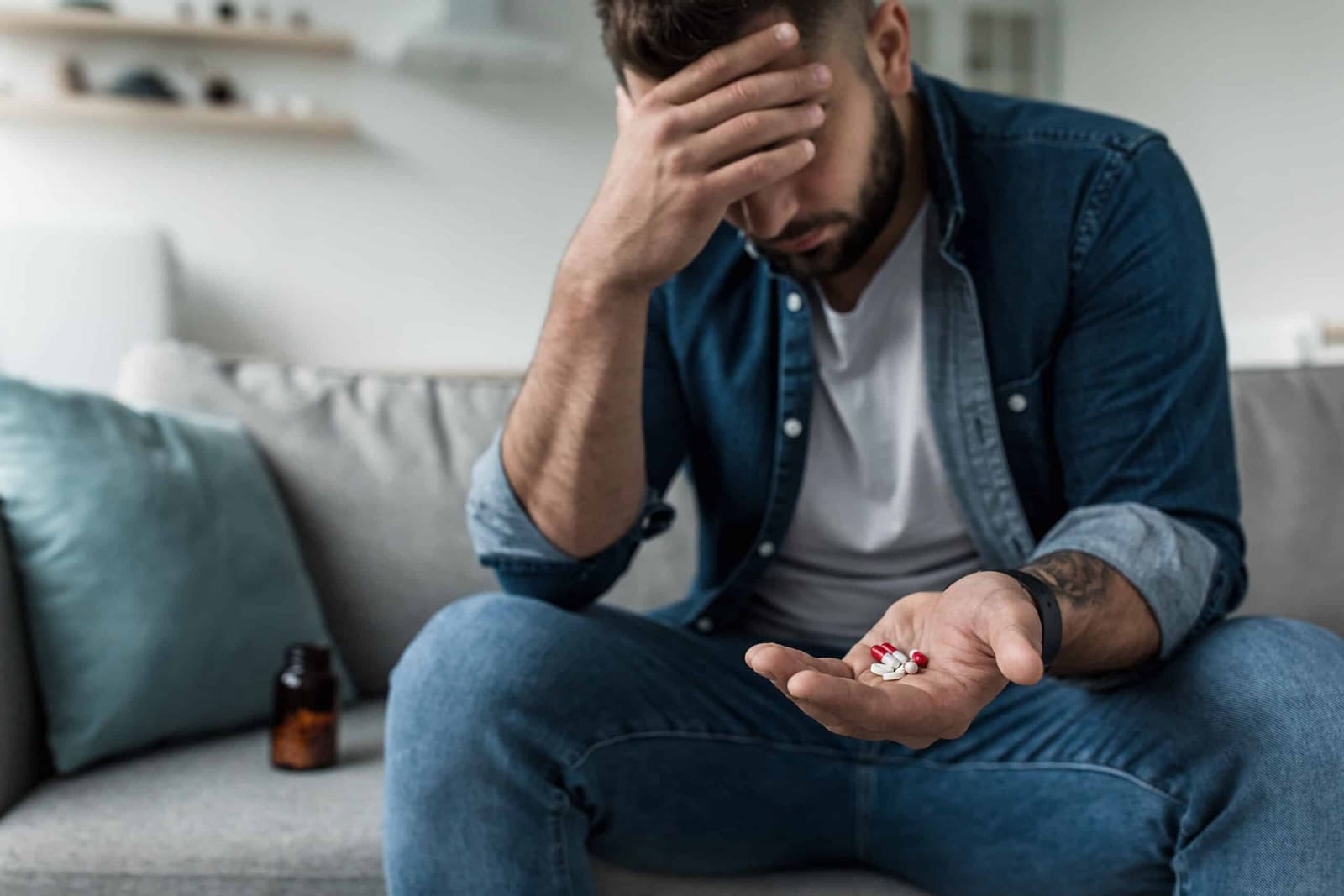 Distressed bearded man sitting on a couch with his head in his hand, holding a palmful of pills beside a medication bottle