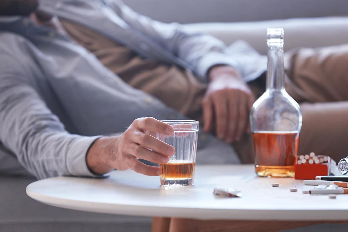 Man slumped on a couch reaching for a glass of liquor beside a bottle, cigarettes, and scattered pills, illustrating alcohol and substance abuse.