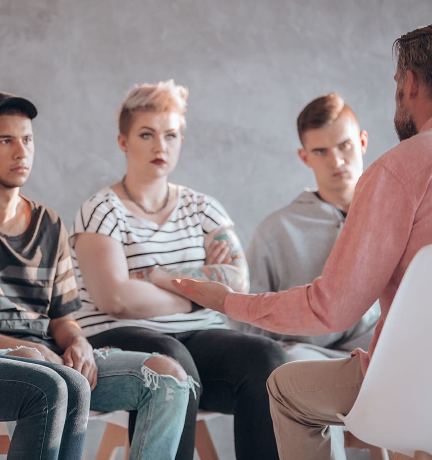 A counselor gestures toward four young adults seated in a circle during a group therapy session