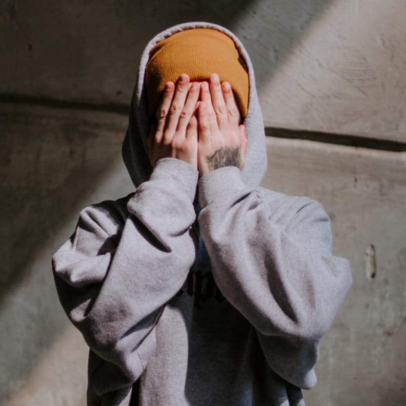 Young person in a gray hoodie covering their face with both hands against a concrete wall