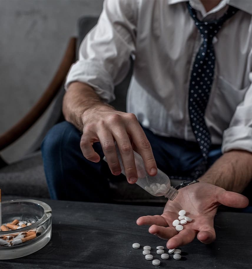 Man in shirt and tie pouring white pills from a bottle into his open hand beside an ashtray of cigarette butts