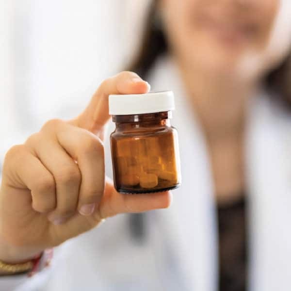 Hand holding a small amber medication bottle with white cap and brown tablets, with a person in a white coat blurred behind