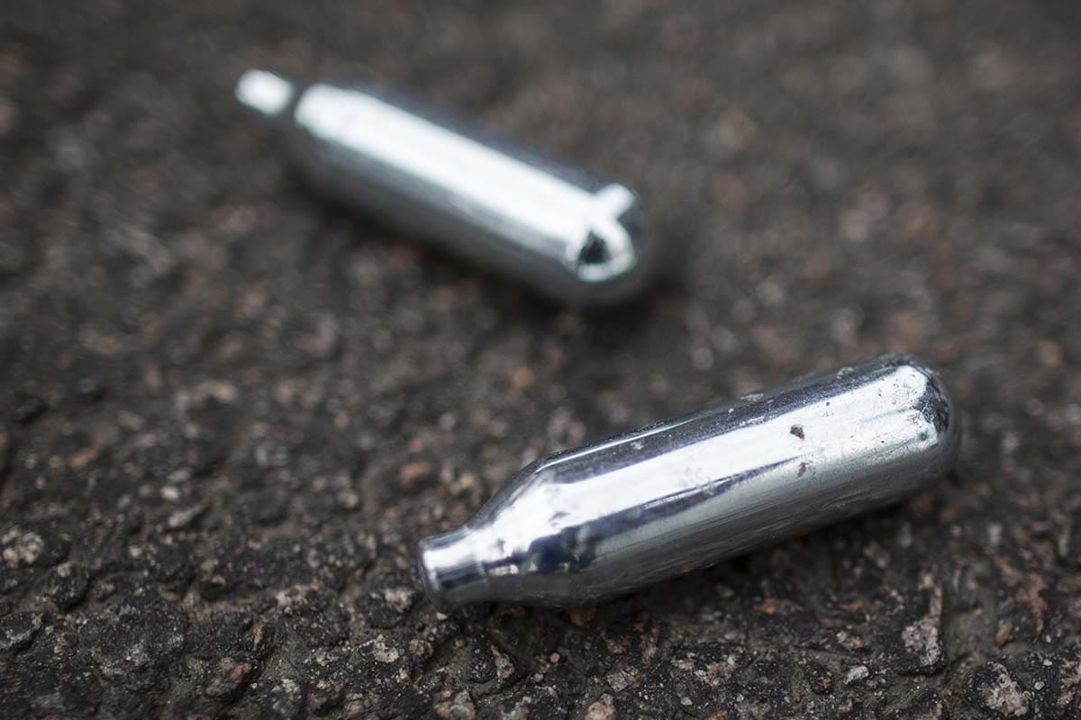 Two used silver nitrous oxide whippet cartridges discarded on dark asphalt pavement