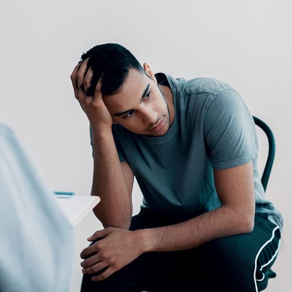 Young man in a gray t-shirt sitting with his head resting on his hand, looking down with a troubled expression.
