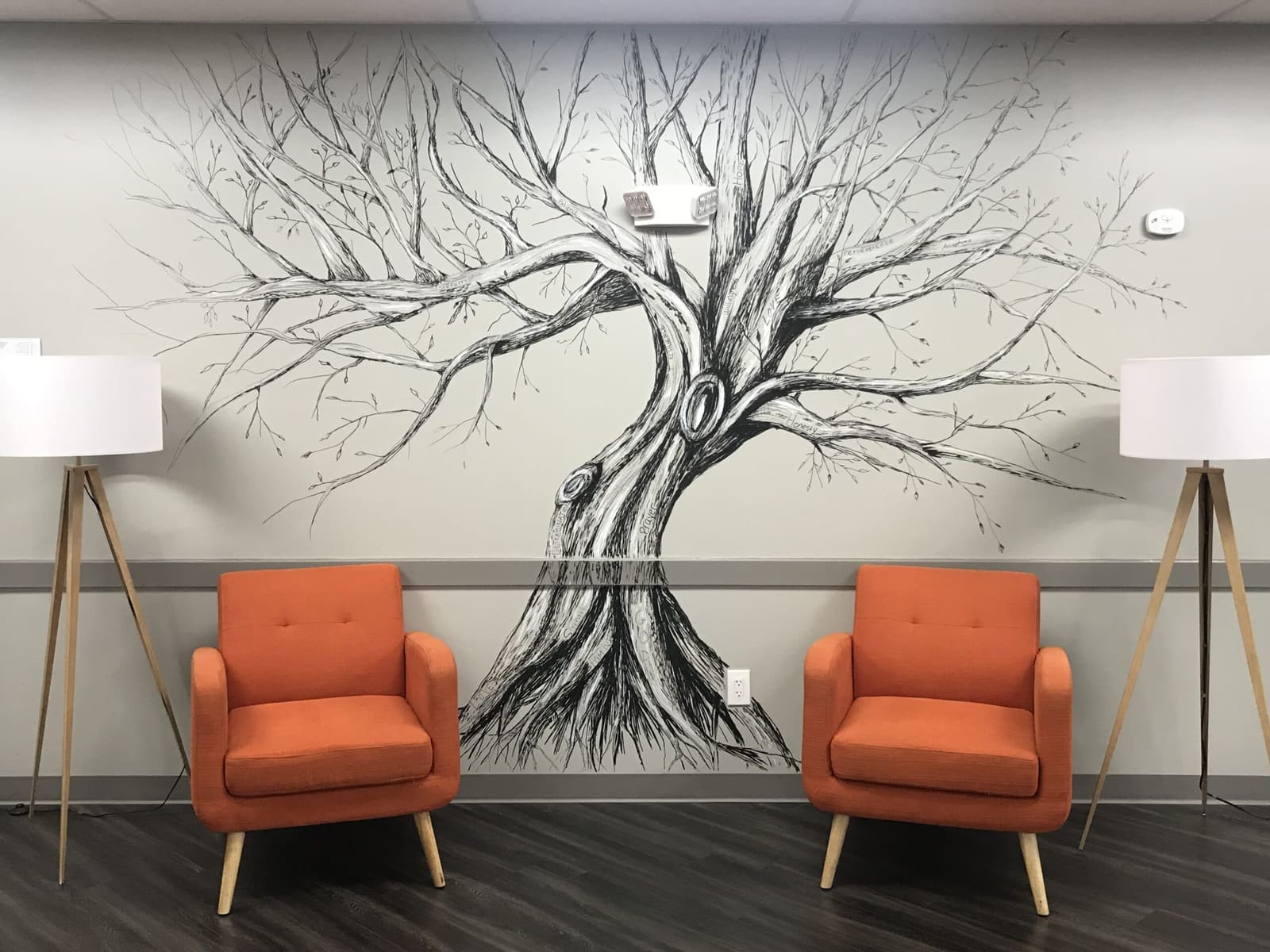 Facility waiting area with a large hand-drawn black-and-white tree mural between two orange armchairs and white tripod floor lamps