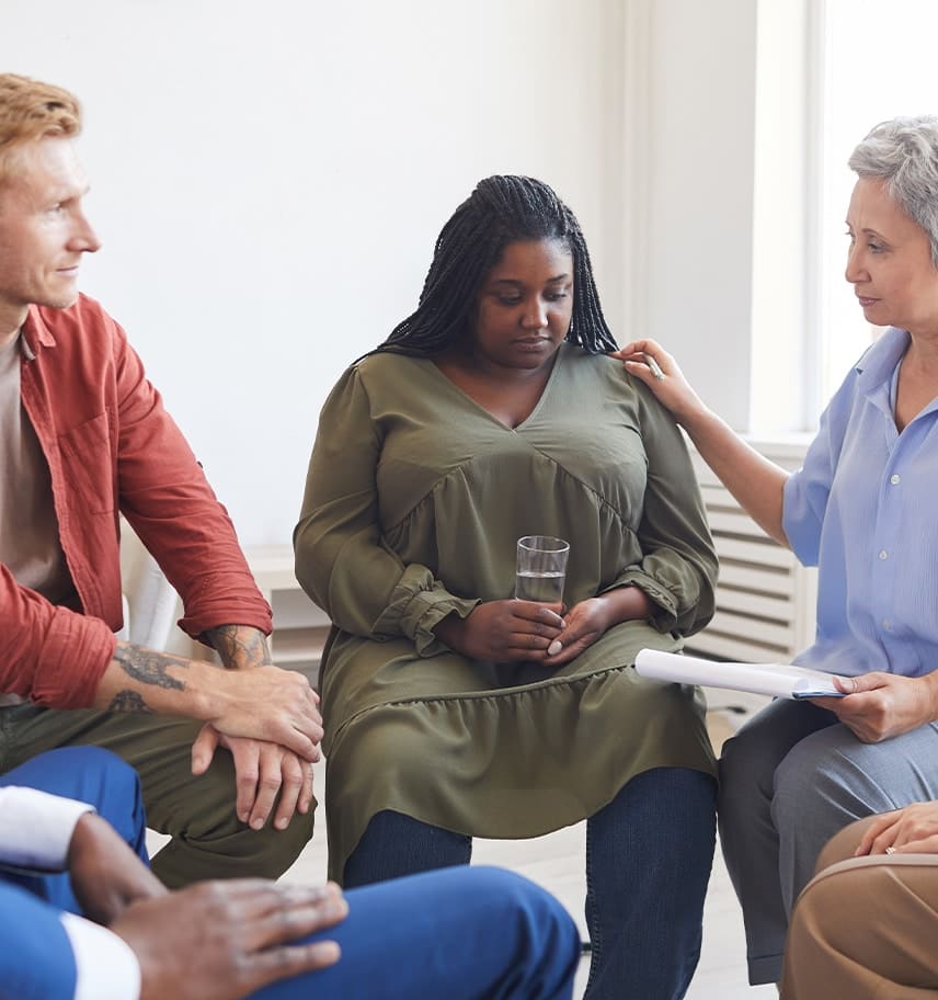 Clinician resting a reassuring hand on a woman's shoulder during a diverse group therapy session