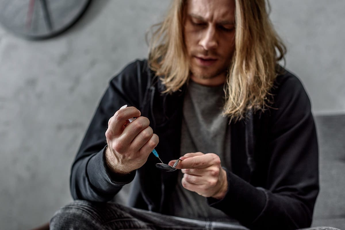 A man heating a spoon to prepare a heroin dose beside a syringe in dim lighting
