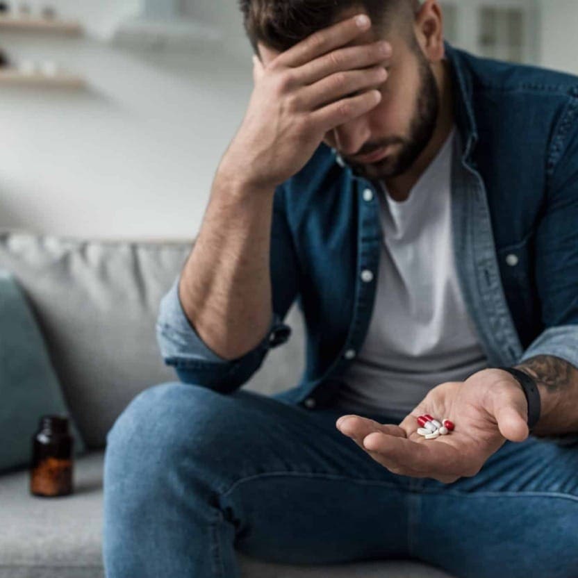 Distressed man sitting on a couch with his head in one hand while holding a palm of pills next to a prescription bottle