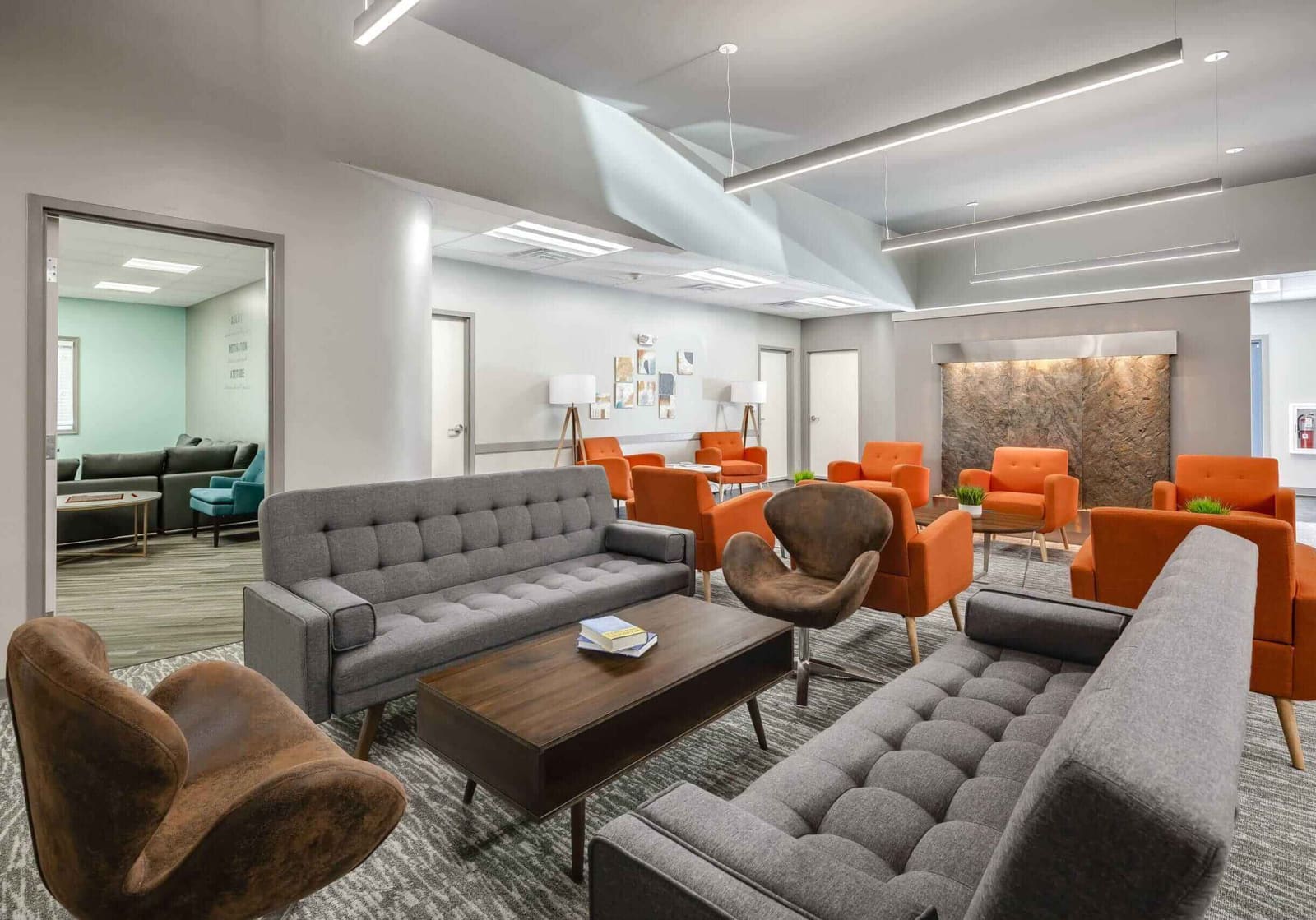 Modern treatment center lounge with gray sofas, leather chairs, and orange accent armchairs in a bright open common area