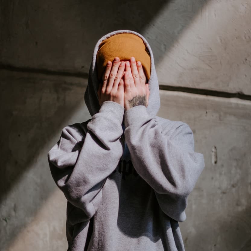 Young person in a grey hoodie and orange beanie covering their face with both hands against a concrete wall