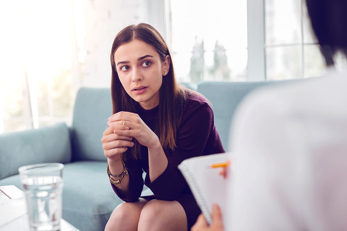 A young woman sits on a couch with clasped hands, looking concerned while talking with a therapist who holds a notepad during a counseling session.