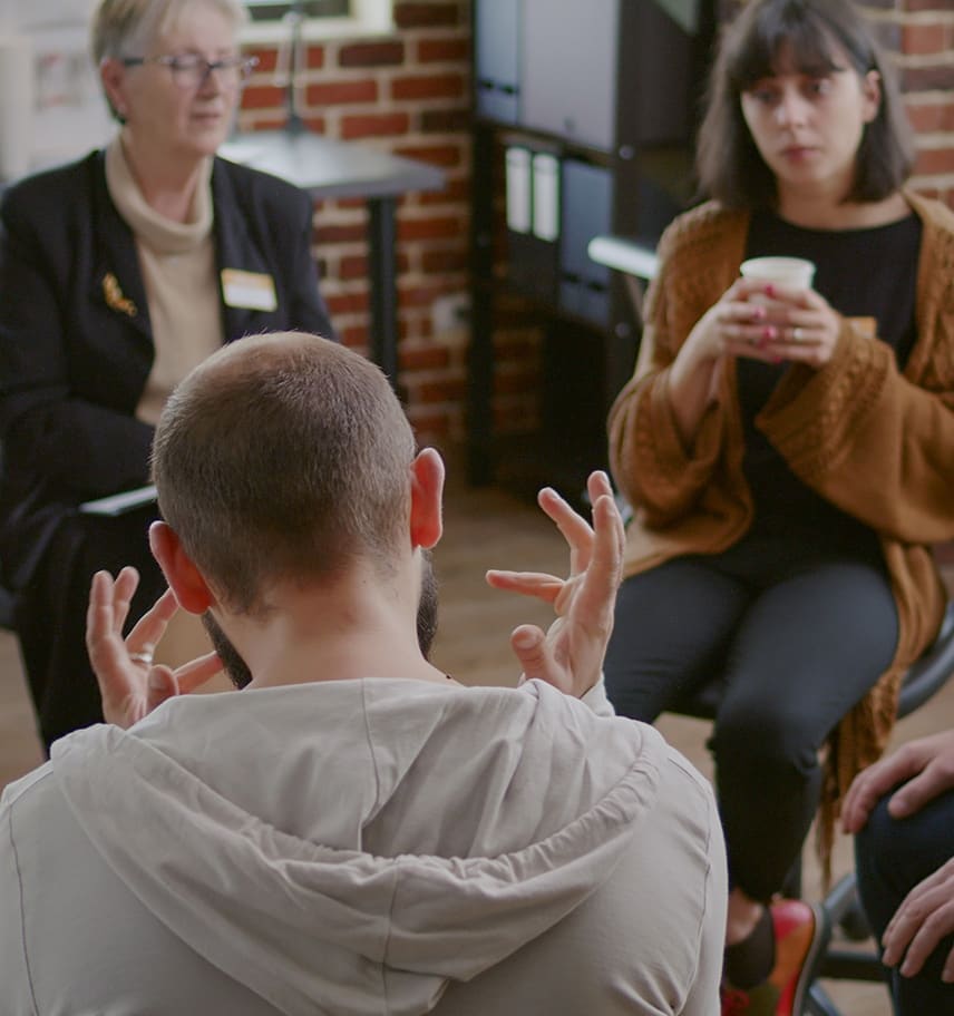 Group therapy session in a brick-walled room where a man gestures while two women listen attentively