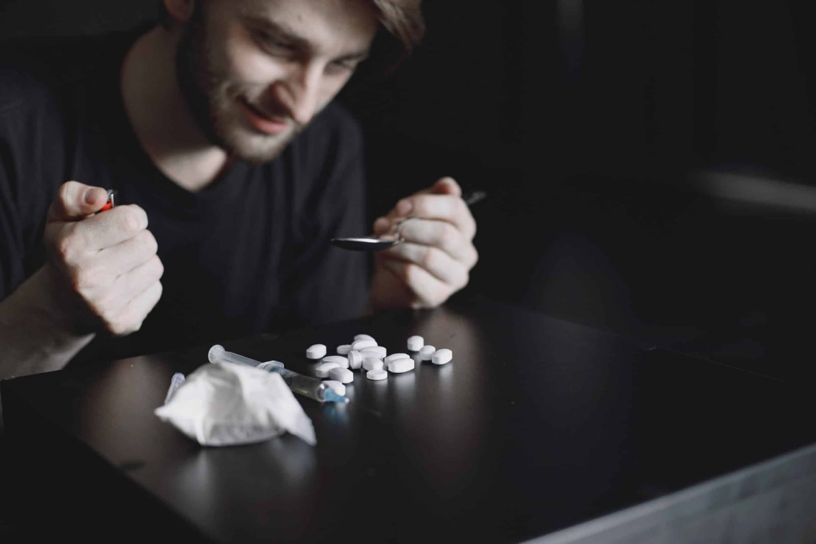 A young man holding a lighter and spoon over a table scattered with white pills, a syringe, and powder in dim light
