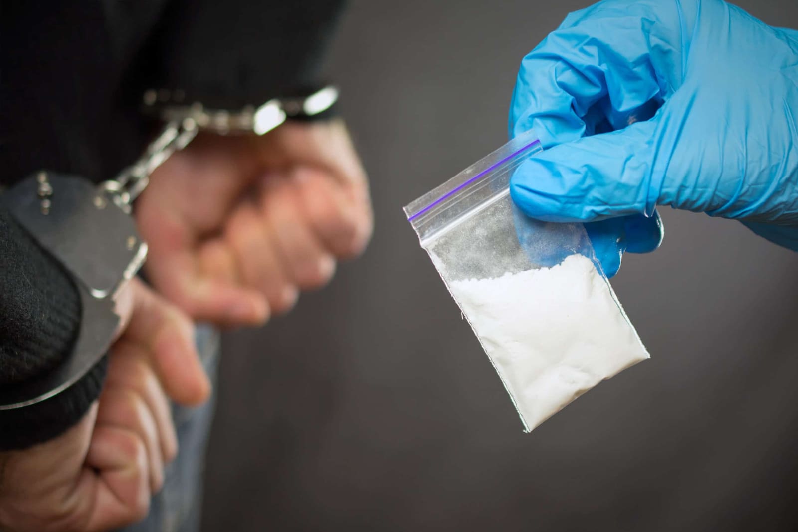 A person handcuffed behind their back as a gloved hand holds a bag of seized white powder