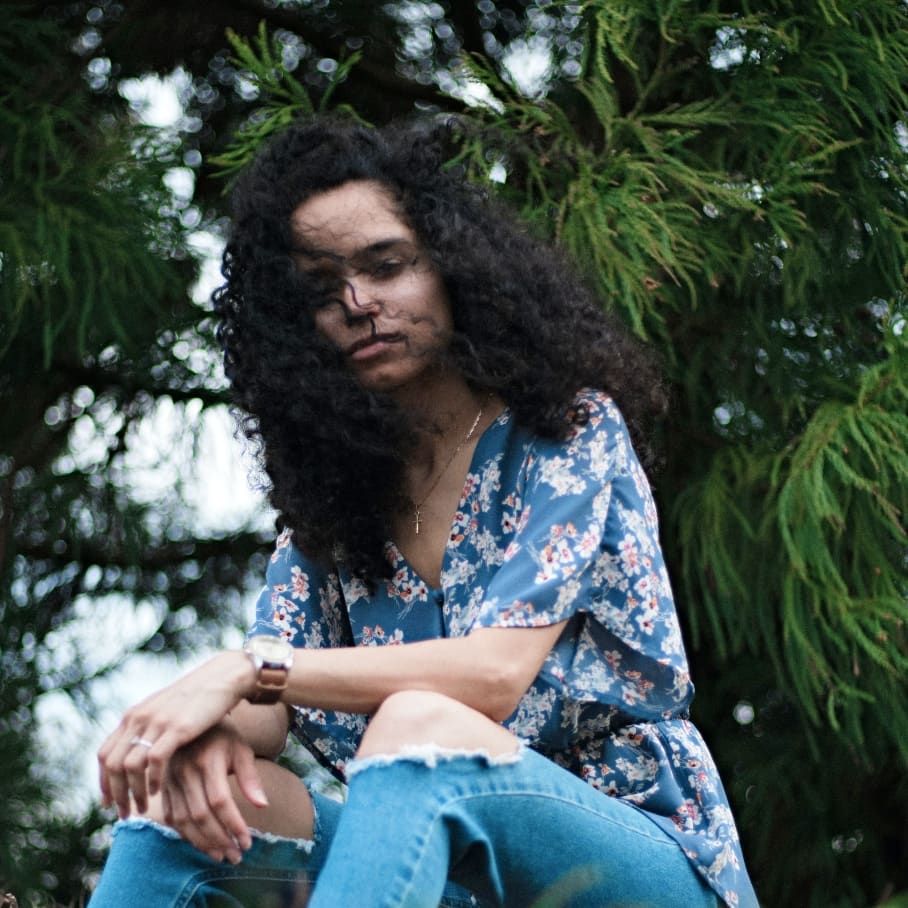 Young woman with long curly hair sitting outdoors against an evergreen tree, head down with a contemplative, withdrawn expression