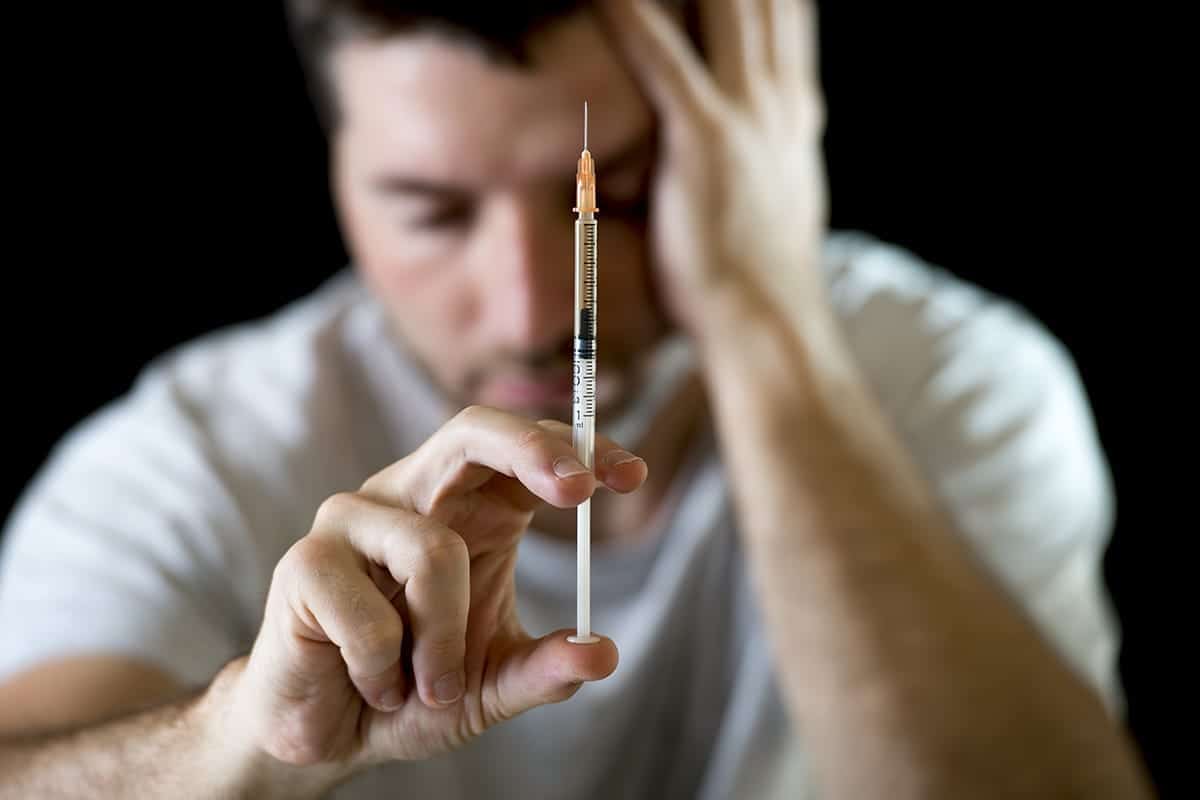 Distressed man in a white shirt holding his head while holding a heroin syringe against a black background