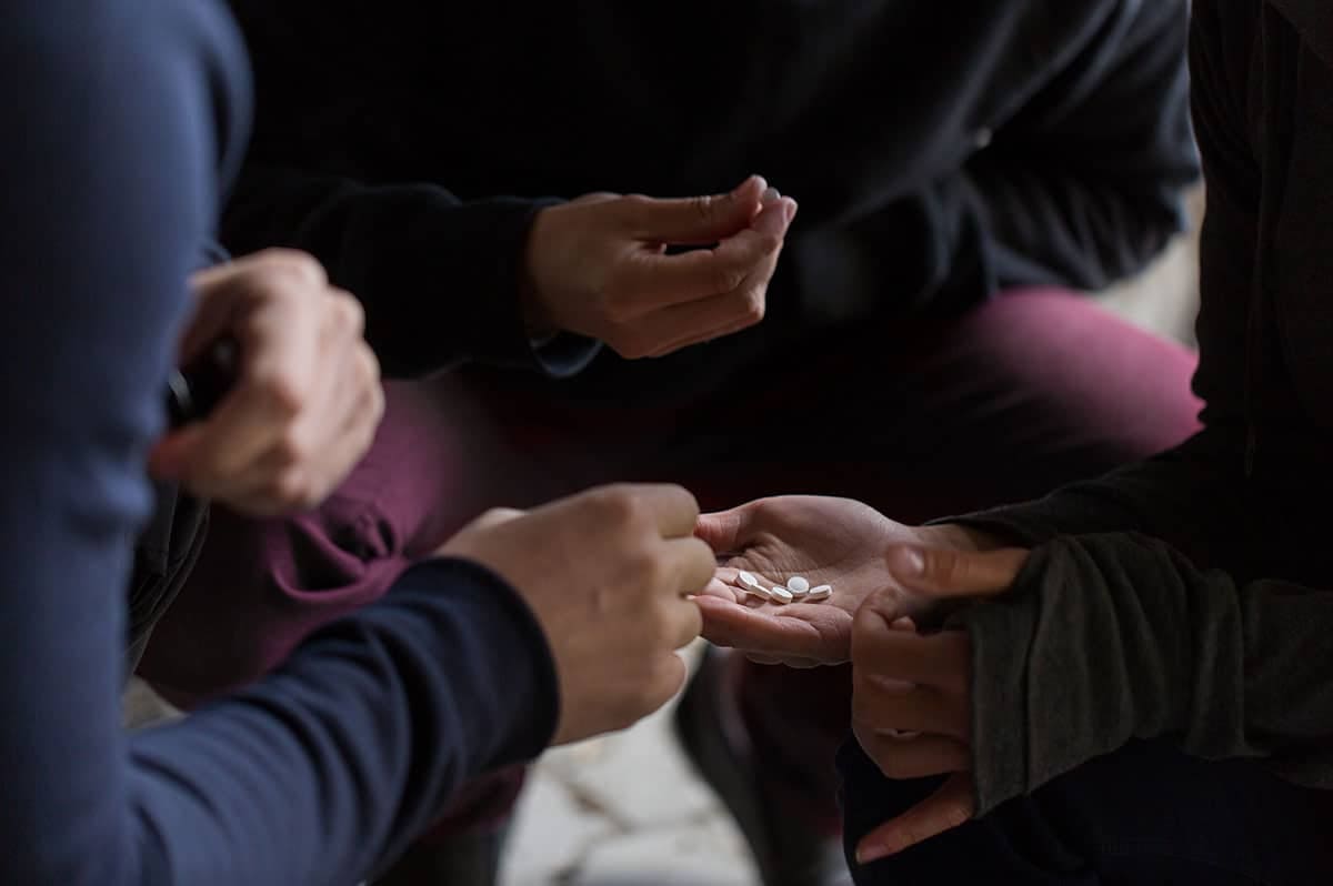 Close-up of hands passing two small white pills between people in dim lighting