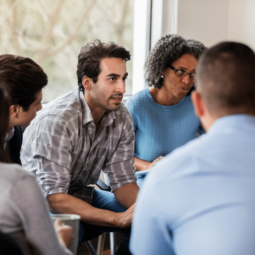 Adults seated in a circle listening to one another during a group therapy session in a sunlit room
