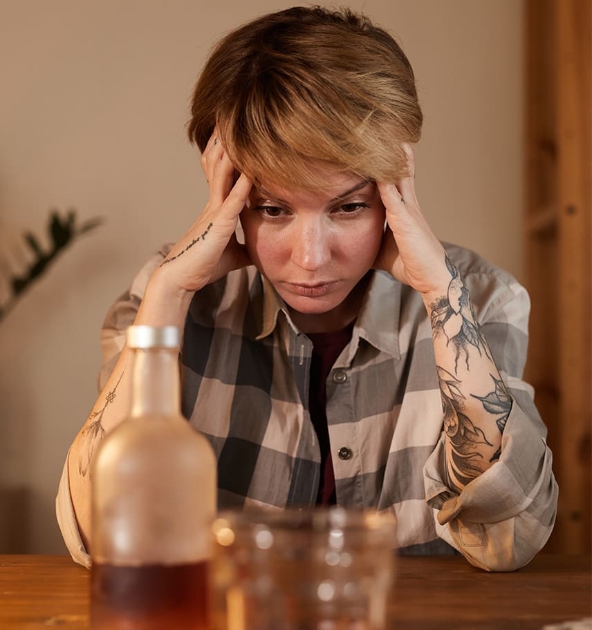 Distressed young woman holding her head at a table with a liquor bottle and glass in front of her