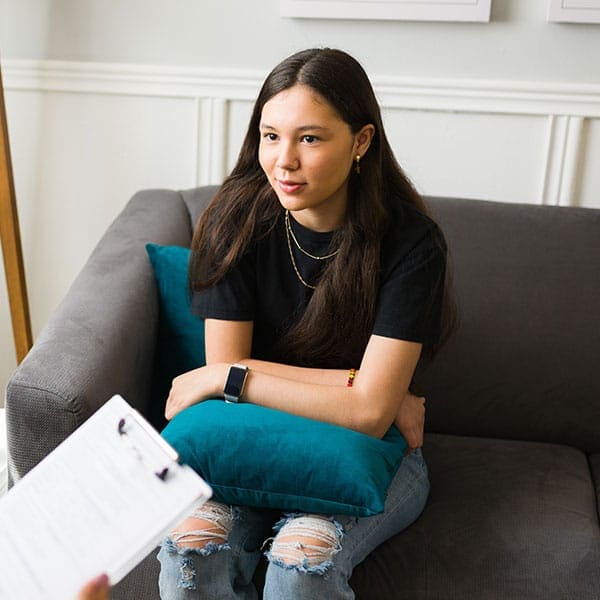 A young woman sits on a gray sofa holding a teal pillow during a therapy session as a clinician takes notes on a clipboard