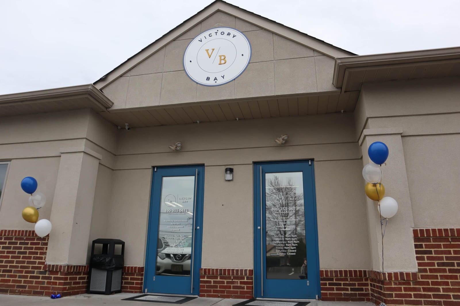 Front entrance of the Victory Bay treatment center, a tan stucco and brick building with teal glass double doors and the round Victory Bay logo on the gable.