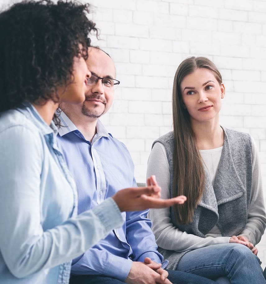 Three adults in a group therapy session, one woman gesturing as she speaks while two others listen
