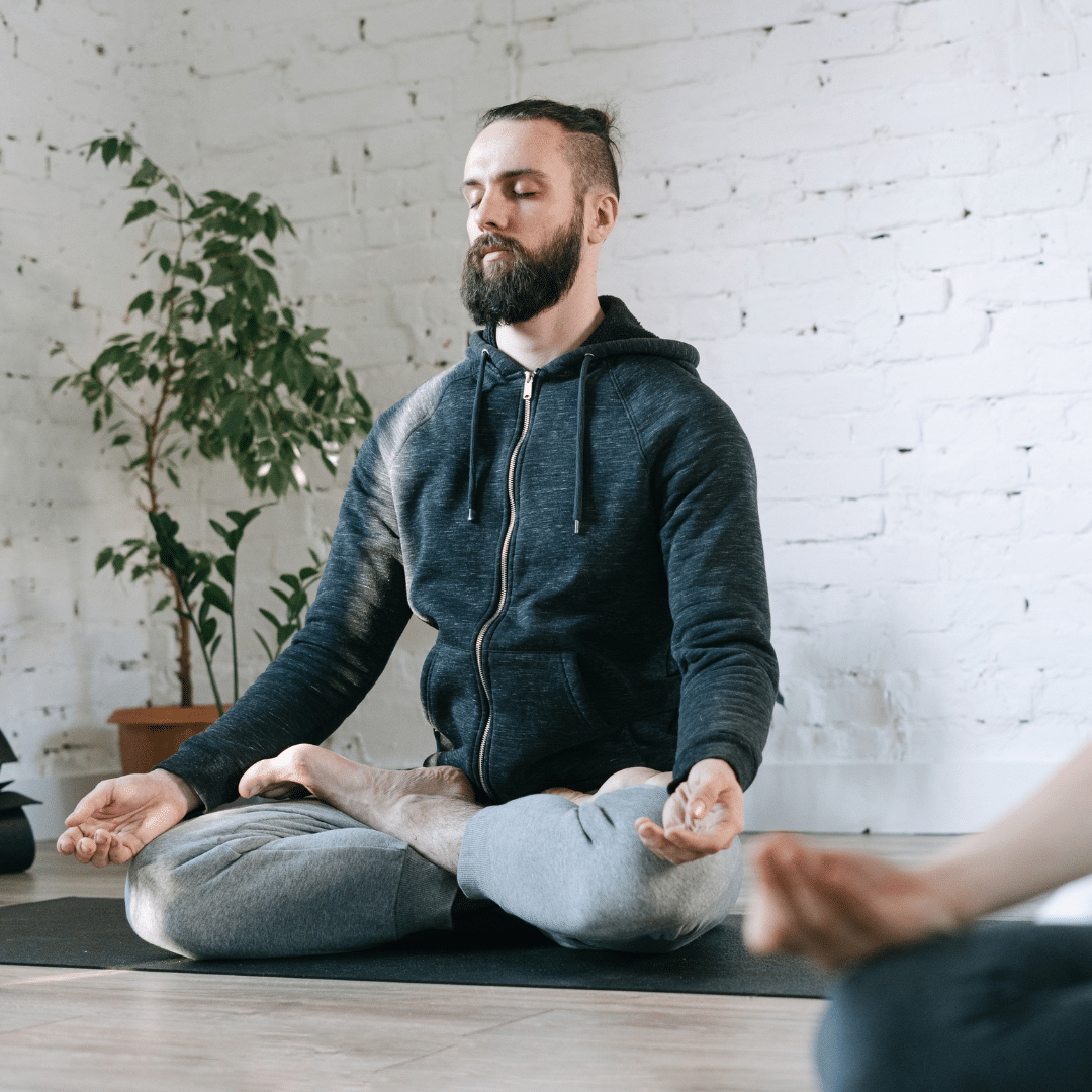 A bearded man sits cross-legged with eyes closed, meditating on the floor of a bright studio during a group mindfulness session.
