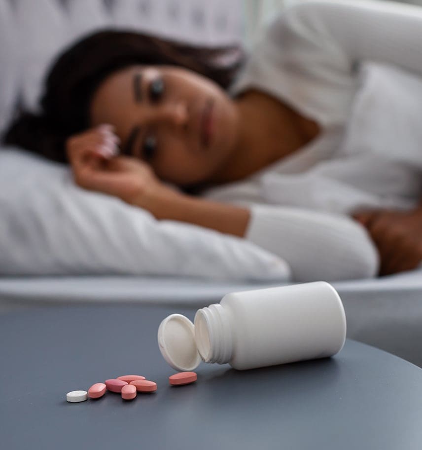 A woman lying in bed beside an overturned prescription bottle with pills spilled across the sheets