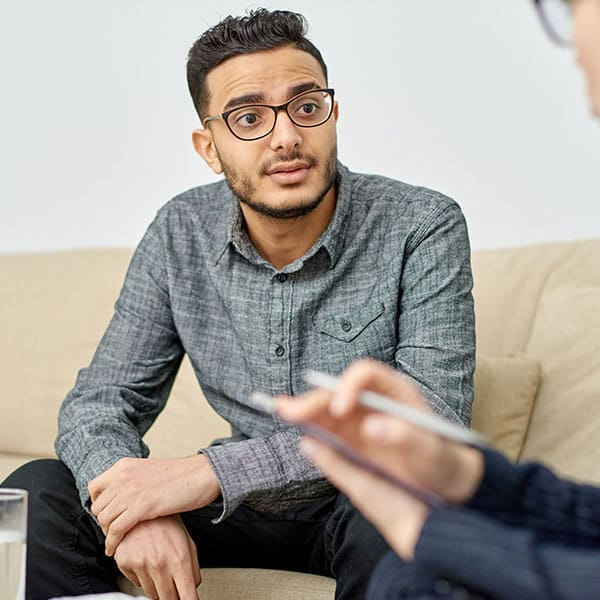 Young man in glasses listening during a one-on-one therapy session as a clinician takes notes on a clipboard