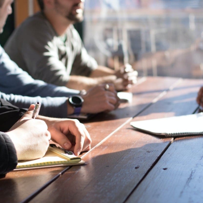 Several adults seated at a wooden table in a sunlit room, one taking notes during a group discussion
