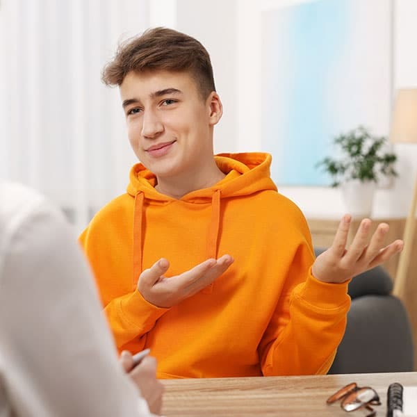 Teenage boy in an orange hoodie gesturing as he talks with a counselor during an indoor therapy session