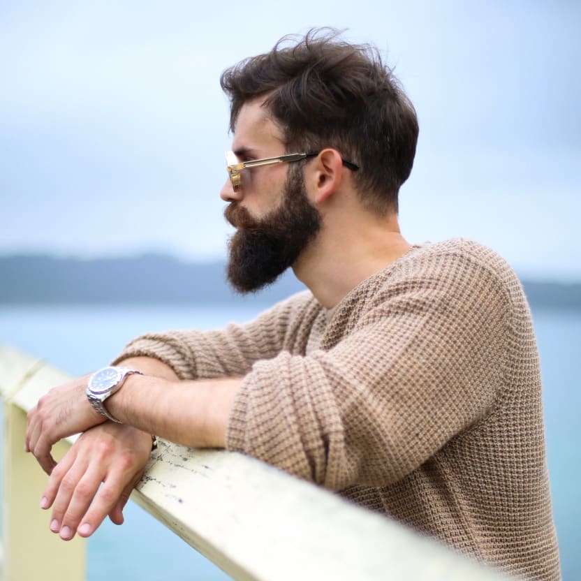 Bearded man in a tan sweater leaning on a railing, looking pensively over calm water