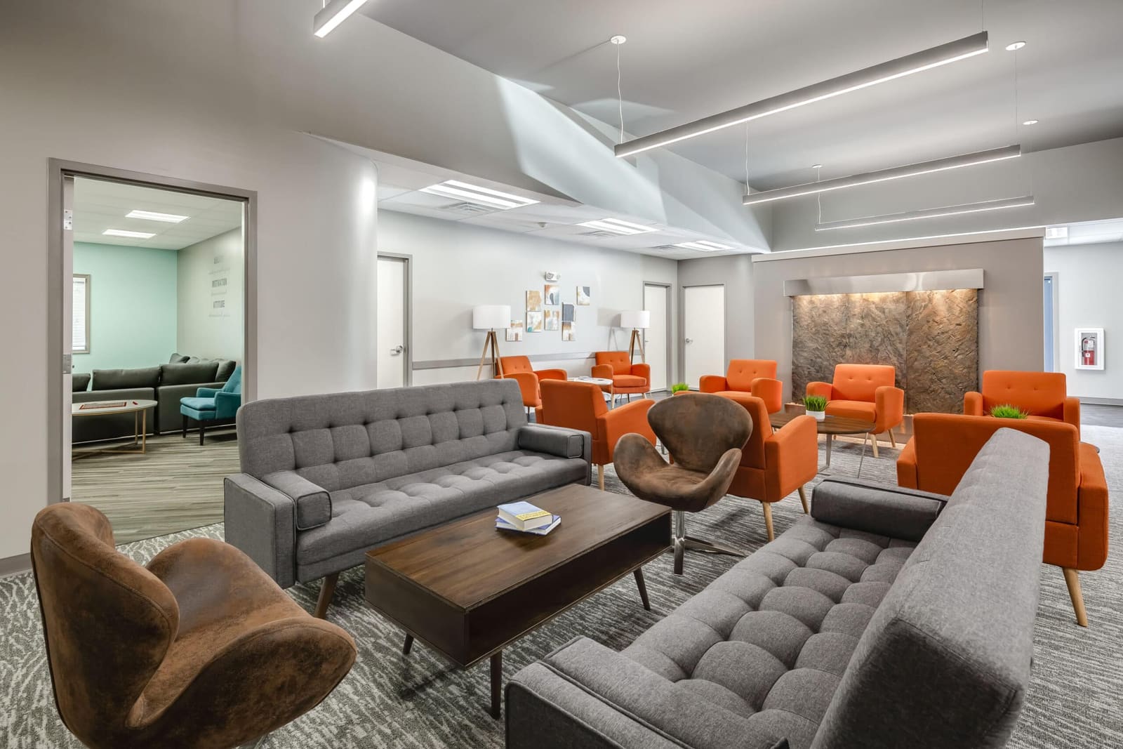 Modern behavioral-health facility waiting room with gray sofas, orange accent chairs, and a wood coffee table on patterned carpet